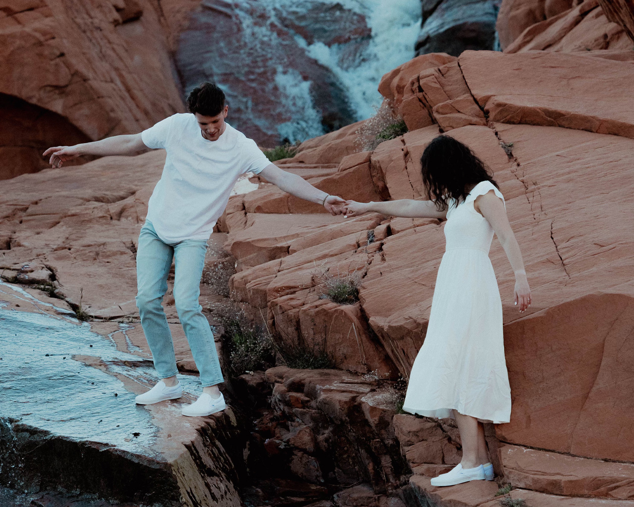 Engagement photo of Bekah and Nick holding hands as they carefully walk across rocks beside a small waterfall in a red rock desert setting