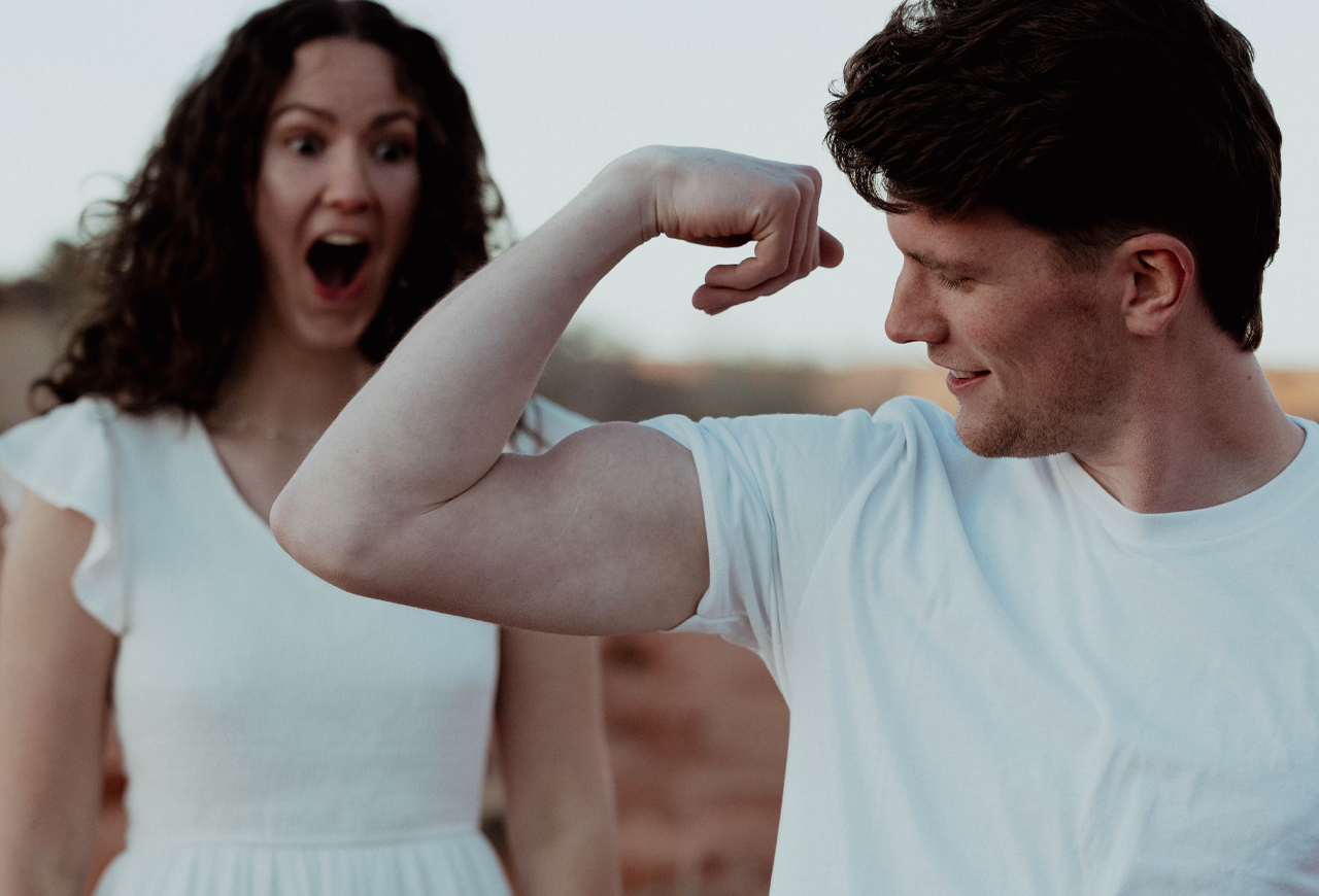 Playful engagement photo of Bekah and Nick in a desert setting, with Nick flexing his arm while Bekah reacts with a surprised smile in the background