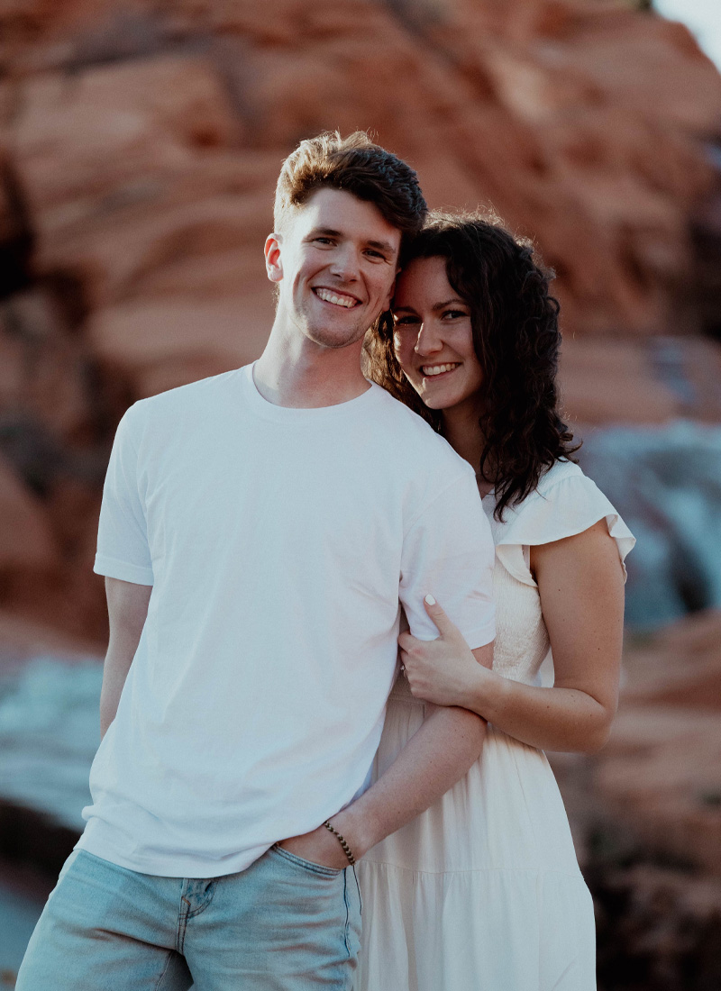 Engagement photo of Bekah and Nick smiling together in a desert setting, with Bekah leaning into Nick as they stand in front of red rock formations